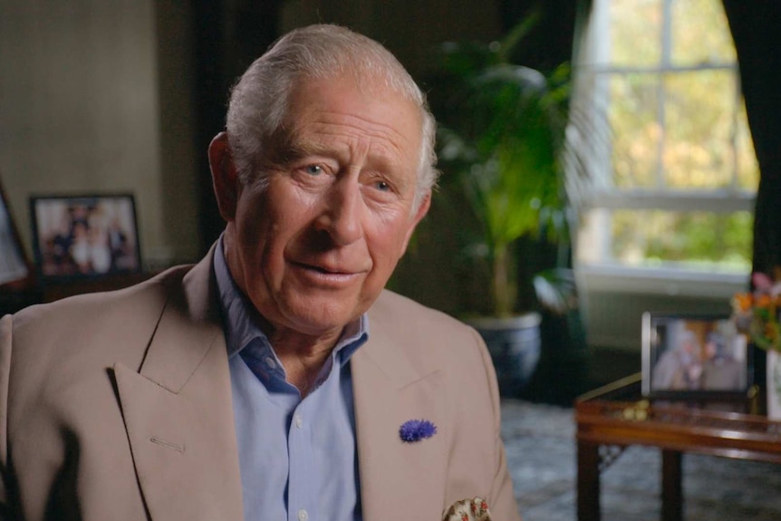 Prince Charles sits in a living room, with family photos in the background.