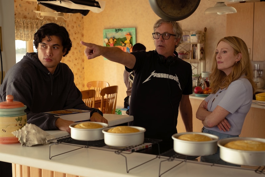 Charles Melton, Todd Haynes and Julianne Moore on set, Haynes pointing into the distance. Cakes are on racks in front of them.