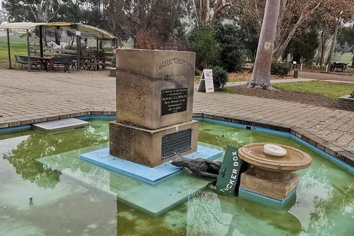 A statue of a dog on a tuckerbox is toppled off its plinth in the middle of a fountain set among a bushy local park.