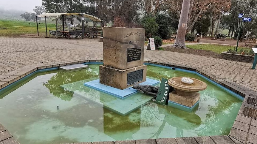 A statue of a dog on a tuckerbox is toppled off its plinth in the middle of a fountain set among a bushy local park.