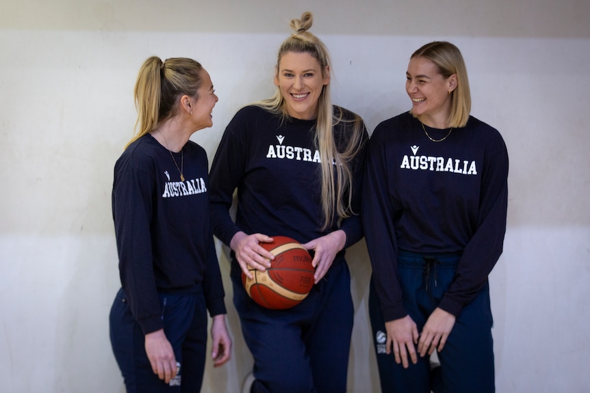 Three women from the Australian Opals basketball squad, in blue shirts that read 'Australia' smile and laugh, holding a ball.