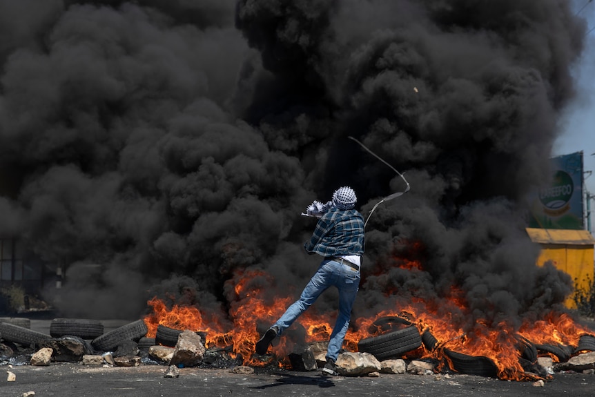 A man fires a slingshot from behind black smoke caused by burning tyres