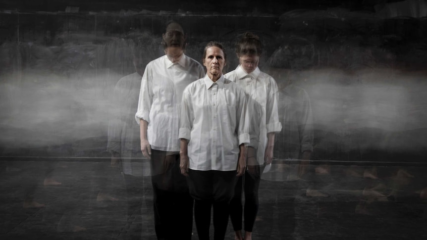 Three women in white shirts and black pants stand in a grey room.