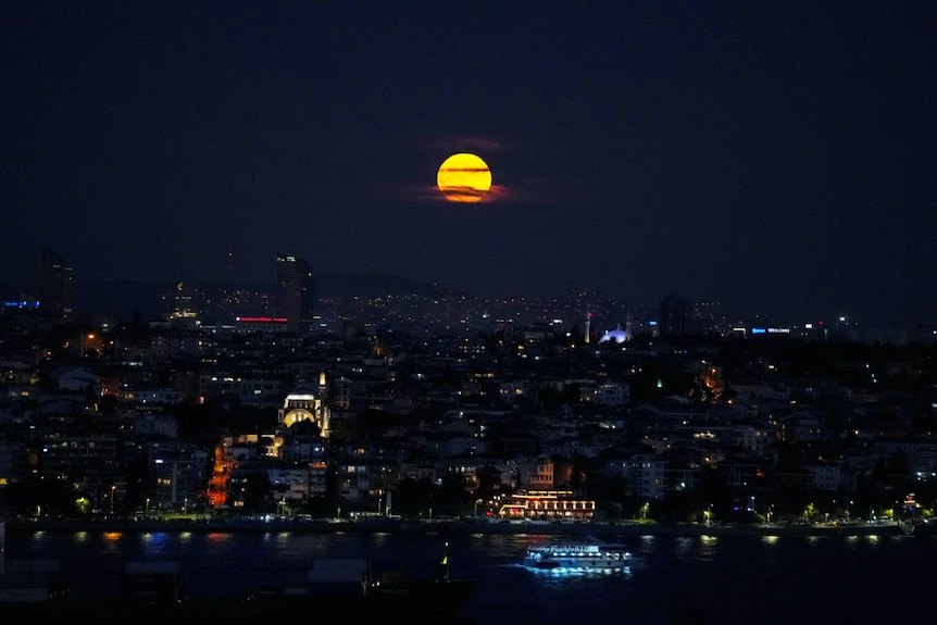 A yellow full moon rises above a city at night
