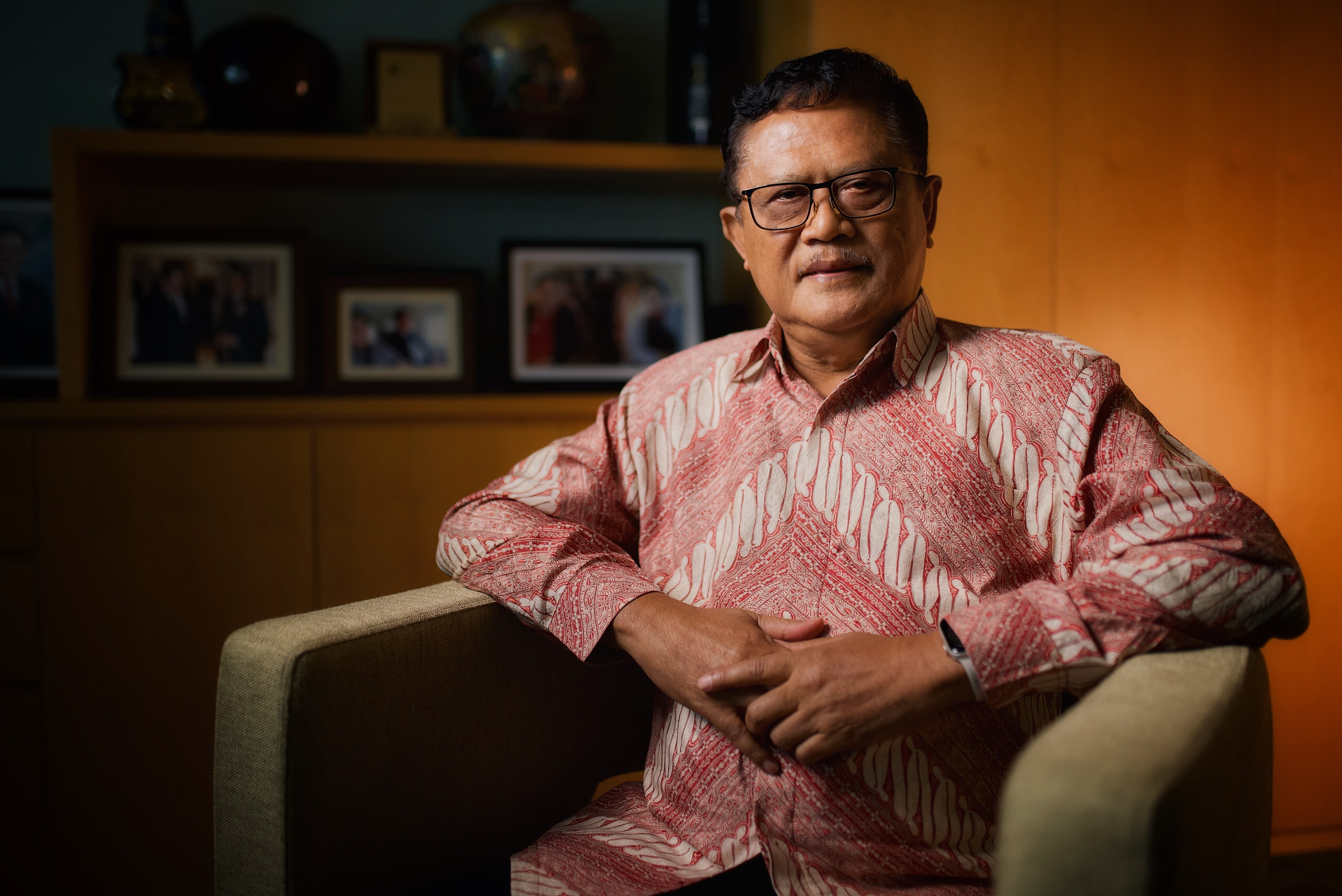 Bachtiar sits with his hands resting on the arms of a chair, folded in towards him, faving the camera with a neutral expression.