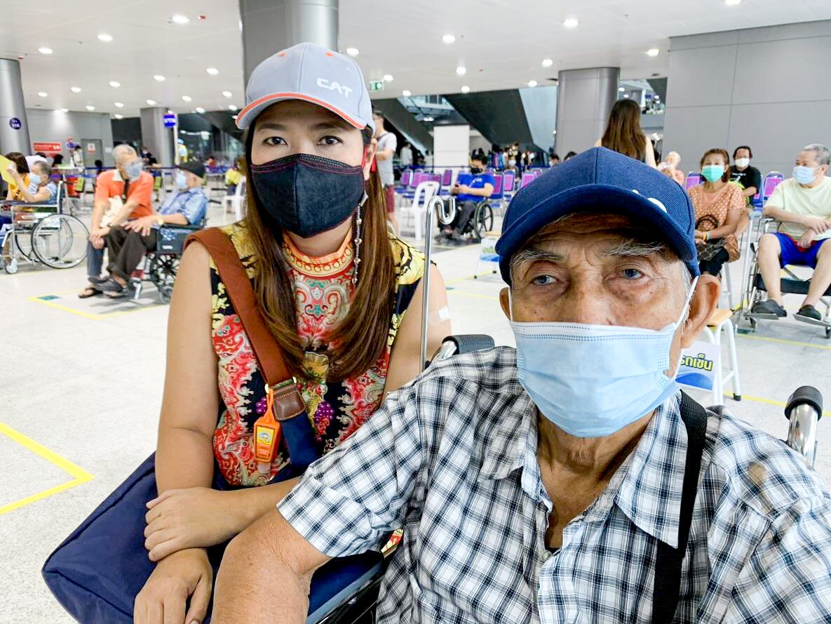 A Thai woman in a face mask crouches next to an elderly man, sitting in a wheelchair