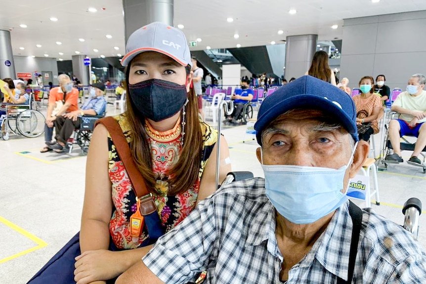 A Thai woman in a face mask crouches next to an elderly man, sitting in a wheelchair