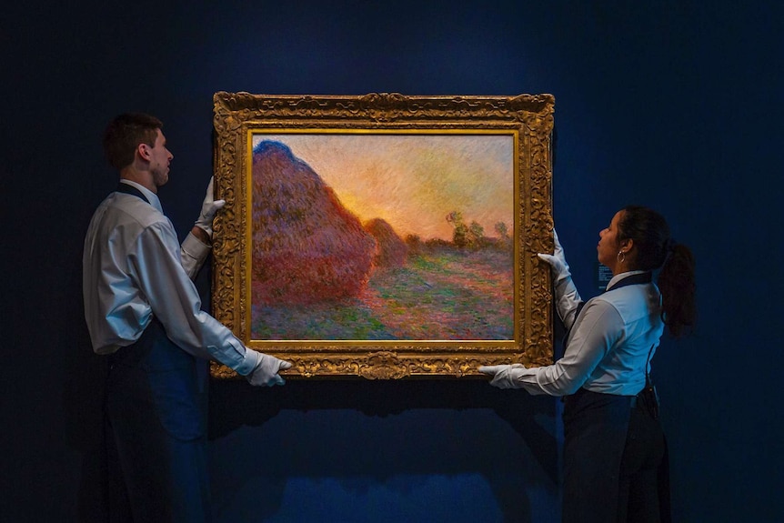 Two gallery attendants, wearing white gloves, lift the gold-framed impressionist painting off a blue wall.