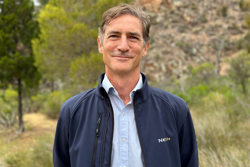 Portrait of brown-haired man wearing blue jacket and blue shirt with trees and hills in the background