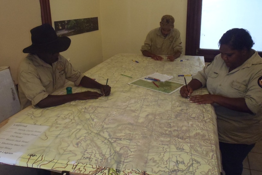 Indigenous rangers sit around a large table tracing elements of a topographic map onto a transparent sheet.