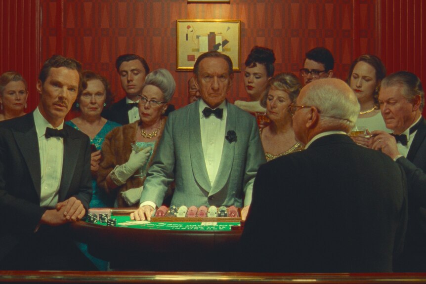 Benedict Cumberbatch and Ben Kingsley stand at a blackjack table surrounded by rich people watching