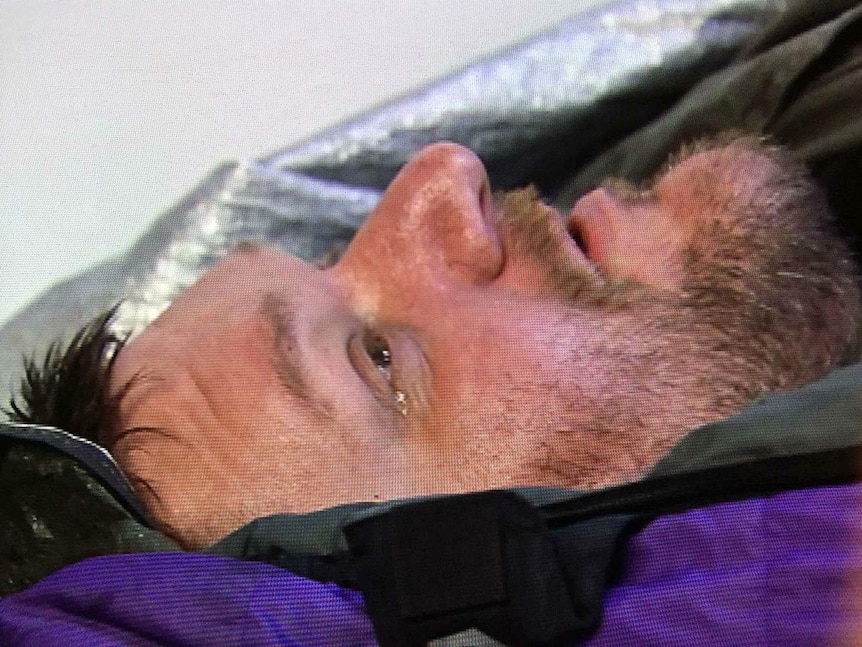 A man lying on an ambulance backboard in a helicopter sheds a tear