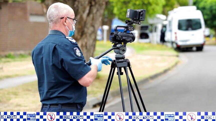 A police officer, wearing a face mask and blue gloves, stands behind a camera on a tripod.