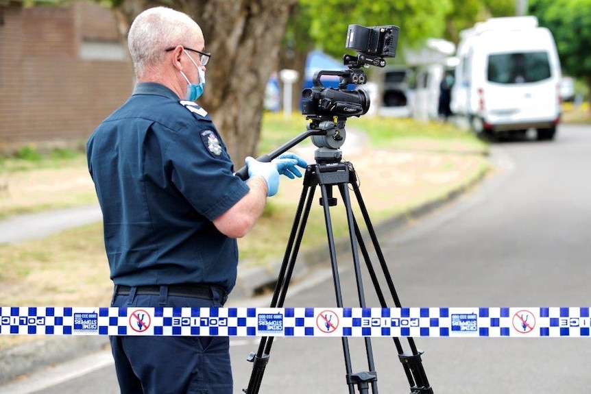 A police officer, wearing a face mask and blue gloves, stands behind a camera on a tripod.