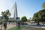 Crowds walk and cycle over a futuristic bridge on a sunny day. 