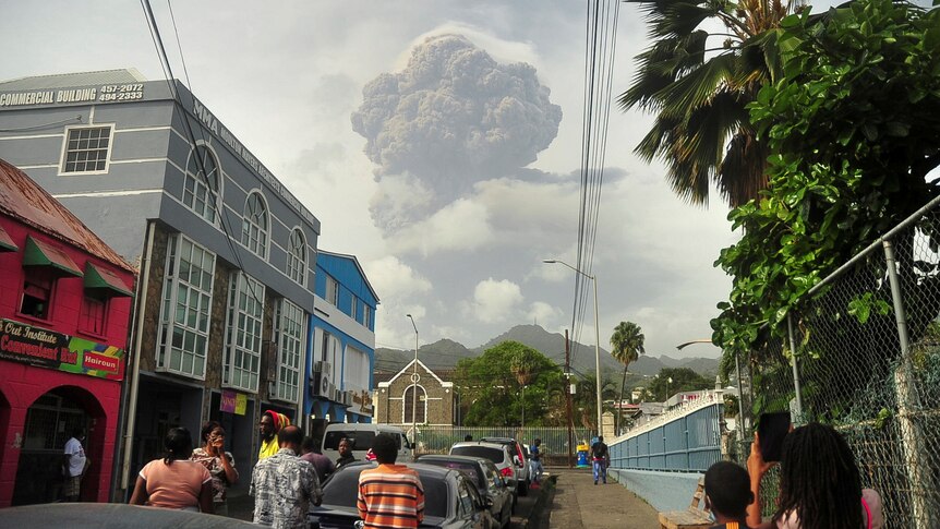 People in a street in a town look at a plume of ash in the distance. 