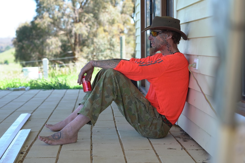Jan Van Der Zon sits on a wooden deck wearing a bright orange top, sunglasses and an akubra.
