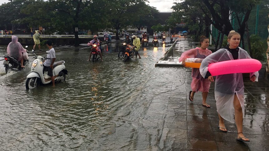 Tourists wear floatation devices while scooter riders pause before crossing a flooded road in Thailand.
