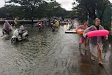 Tourists wear floatation devices while scooter riders pause before crossing a flooded road in Thailand.