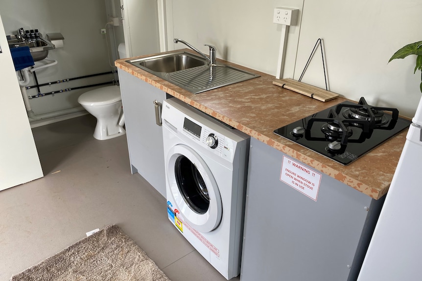 A narrow room showing a small kitchen area with a stove, sink, and washing machine, and a doorway leading into a small bathroom.