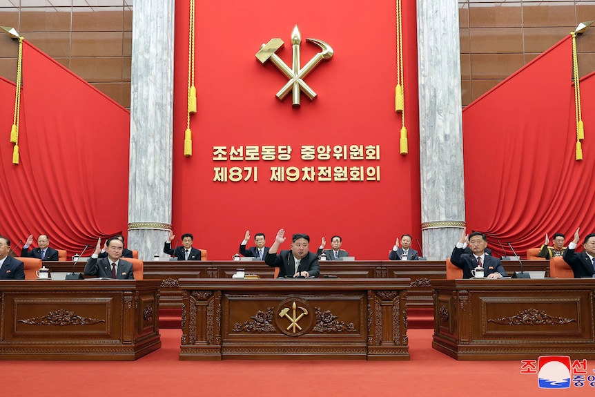 A group of men sit on stage at an official meeting raising their hands. 