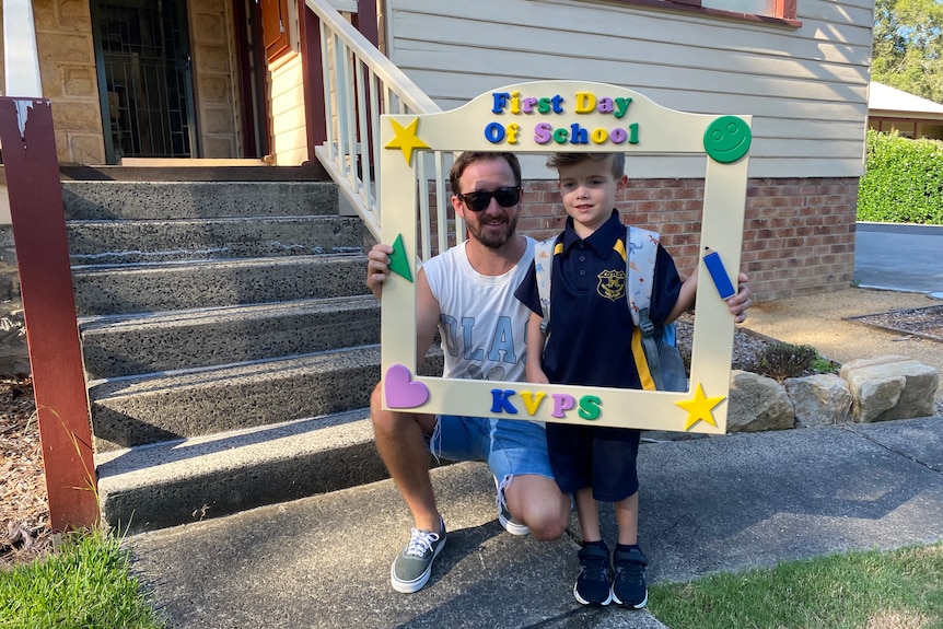 A dad and his son with a first day of school frame around them, taking a photo. 