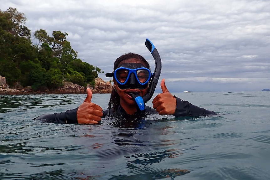 A woman wearing snorkel gear holds her head and body out of the water. She has two thumbs up.