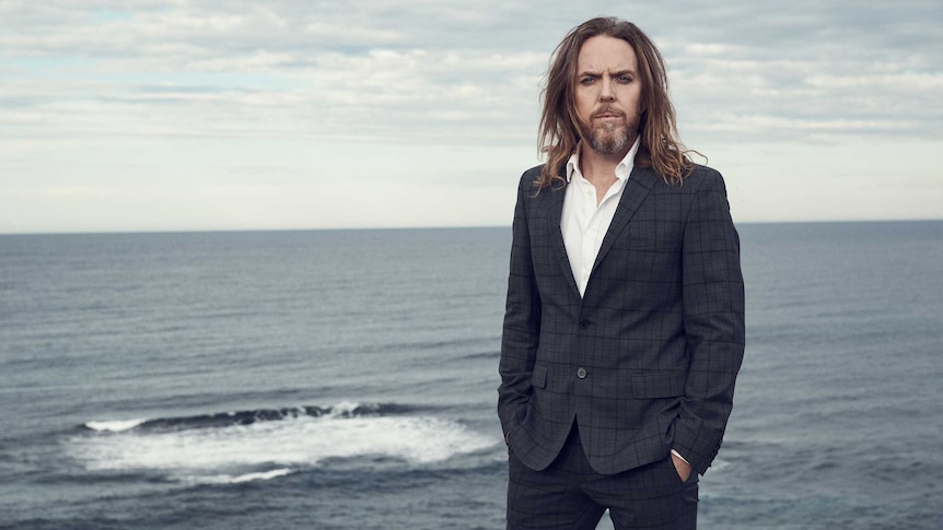 Tim Minchin stands by the ocean wearing a checked suit. He's looking stern and staring at the camera.