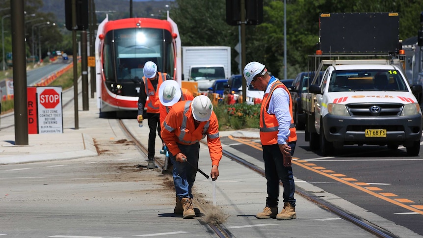 Workers in high-vis jackets clear dirt from the light rail tracks. A light rail vehicle can be seen behind them.