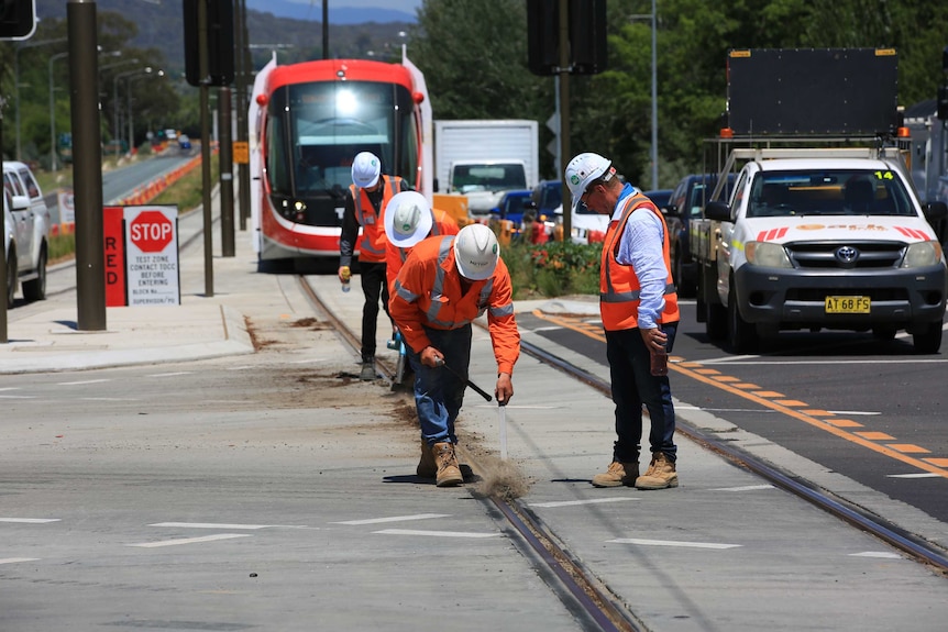 Workers in high-vis jackets clear dirt from the light rail tracks. A light rail vehicle can be seen behind them.