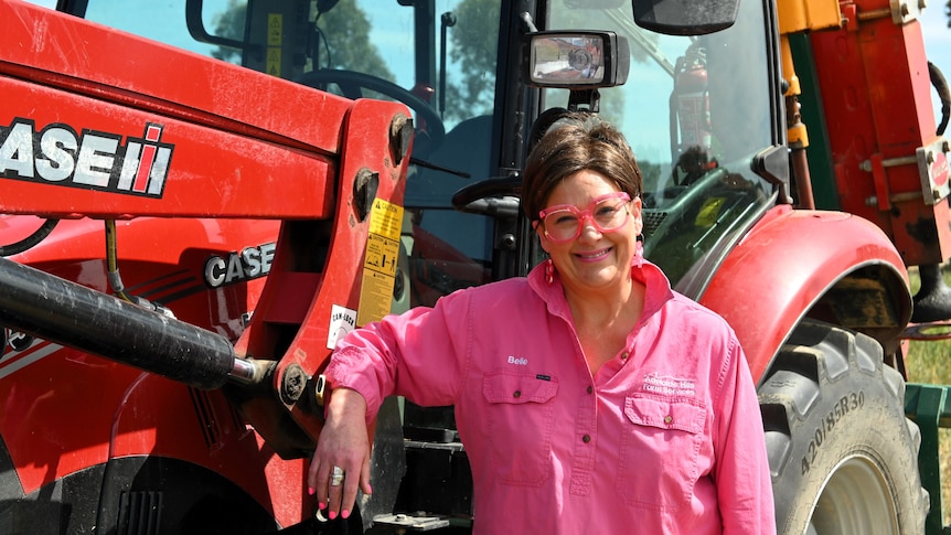 A smiling woman in a pink shirt and pink glasses stands in front of a tractor.