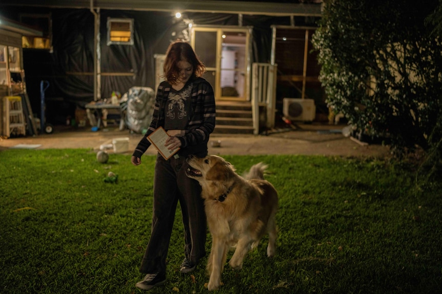 A teenage girl walks in her backyard at night holding a book. She is looking down at her dog, a golden retriever.
