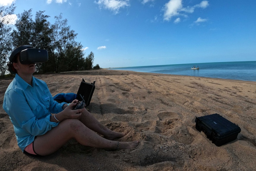 A woman sitting on beach with a remote control, piloting a drone.