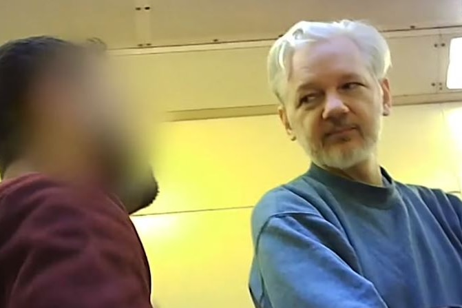 Julian Assange, wearing a blue jumper, talks to a man whose face is blurred.