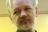 Julian Assange, wearing a blue jumper, talks to a man whose face is blurred.