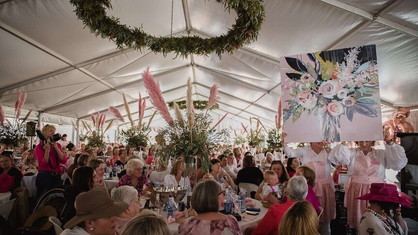 A woman holds up a painting for auction, under a marquee with hundreds of women seated at decorated tables.