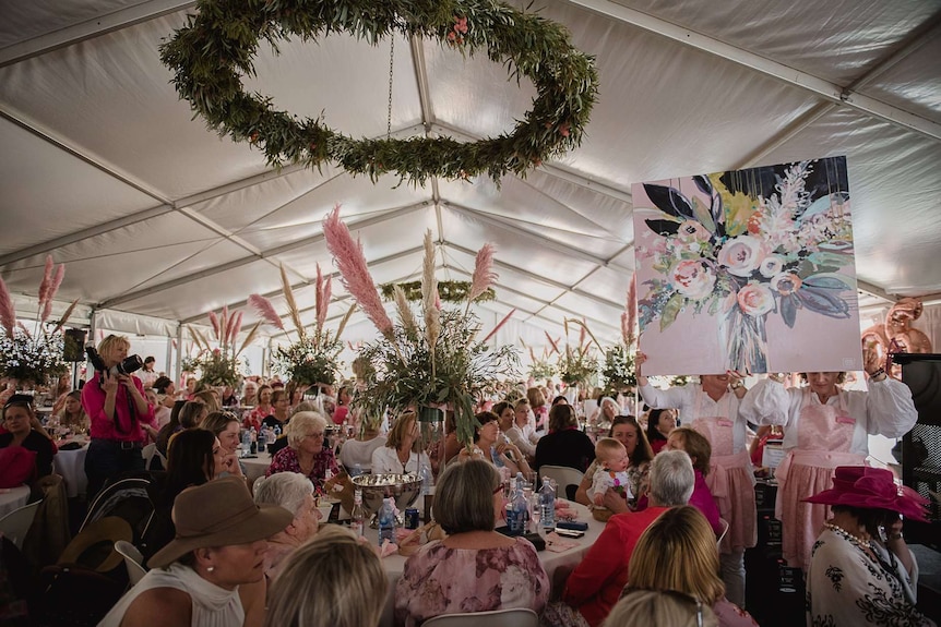 A woman holds up a painting for auction, under a marquee with hundreds of women seated at decorated tables.