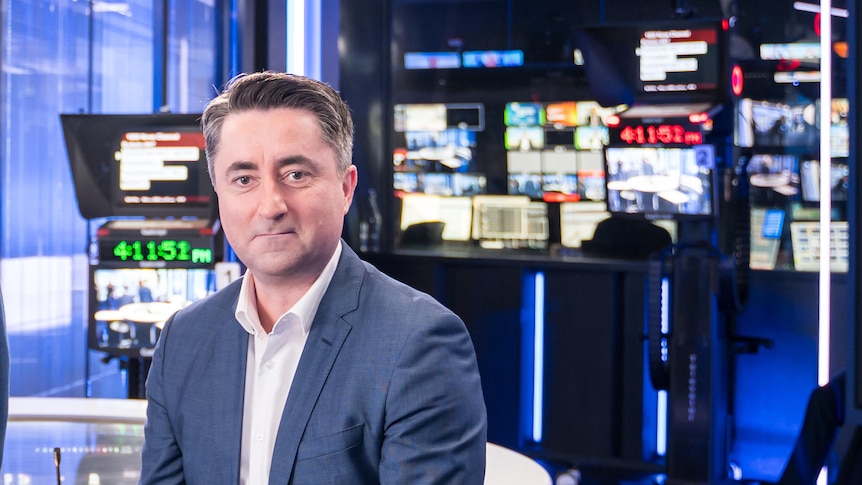 Man sitting at desk in news studio with cameras and monitors in background.