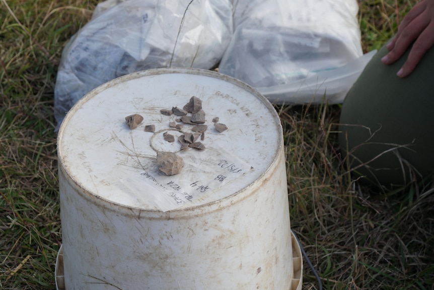 An overturned bucket with a series of small stone artefacts placed on top in a paddock. 
