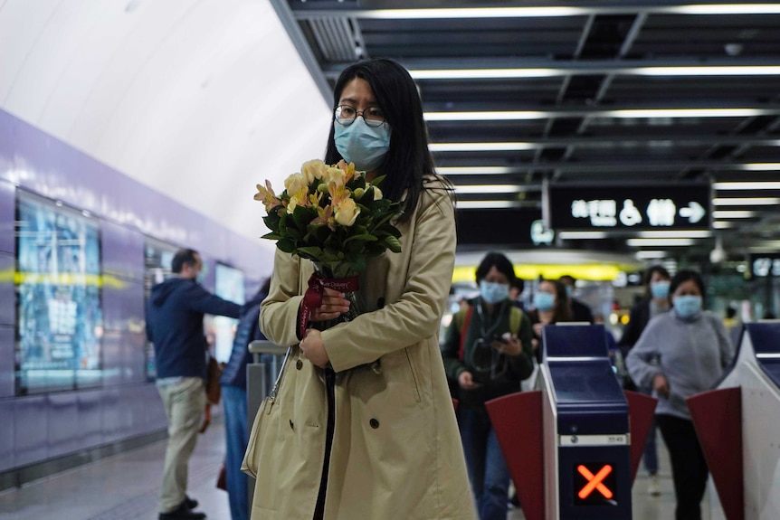 A young woman in an overcoat and wearing a mask holds a bunch of flowers in a subway tunnel