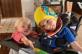 A photo of a smiling young boy in a wheel chair next to a young blonde toddler
