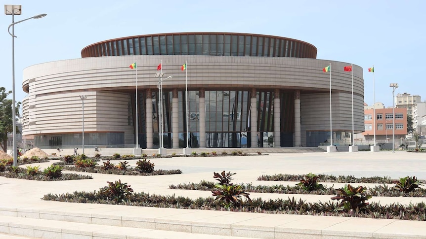 Museum of Black Civilisations building is seen from the road, a beige circular building with a multi-storey glass entrance.