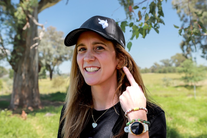 Woman wearing black cap and t-shirt smiles, pointing to earing while standing outside on farm
