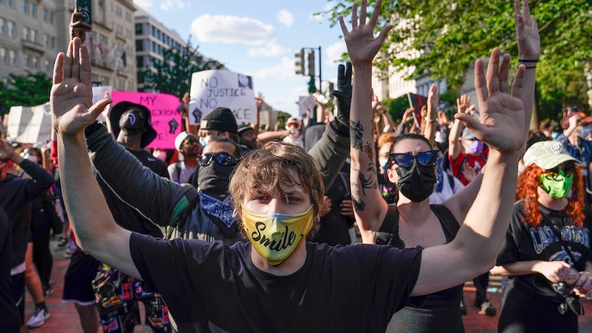 A group of protesters wearing face masks lift their arms on a sunny day in suburban street.