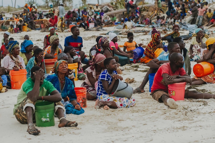 Hundreds of African people sitting on a beach wearing very colourful clothes as they wait with containers for food supplies