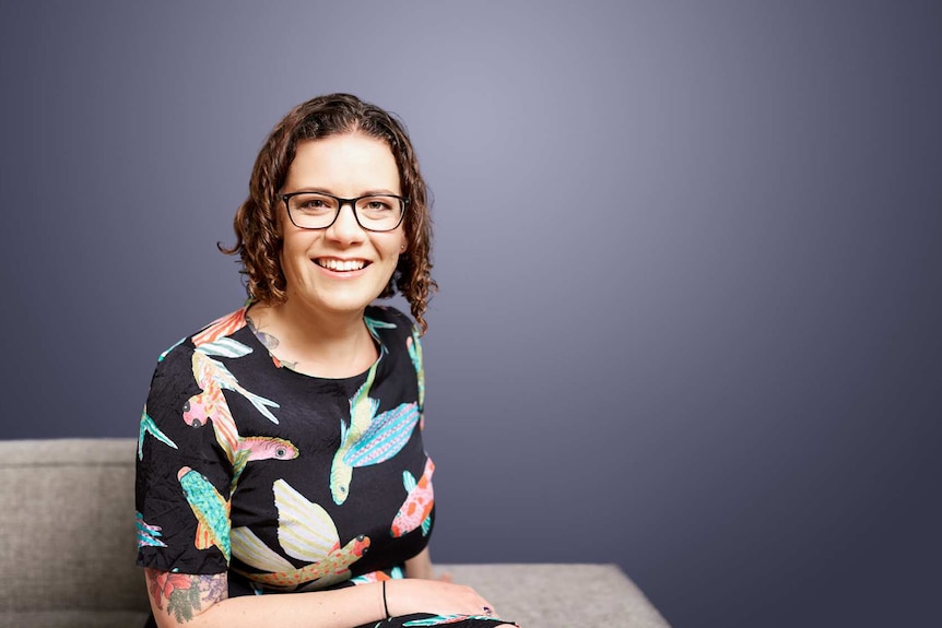 Sarah Jefford smiles at the camera. She's sitting on a couch, wearing a dress with birds printed.