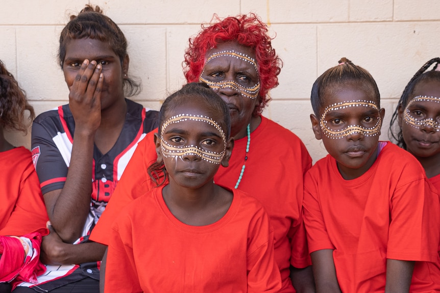 Children in red sit next to each other against a white wall.