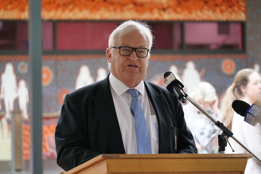 A man with white hair with a blue checkered tie speaking at a lectern