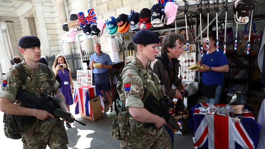 Two heavily armed soldiers walk past tourist souvenir stalls decorated with Union Jacks and selling hats in London.