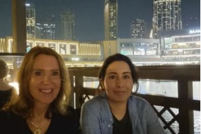 Two women sit on a balcony with skyscrapers visible in the distance behind them.